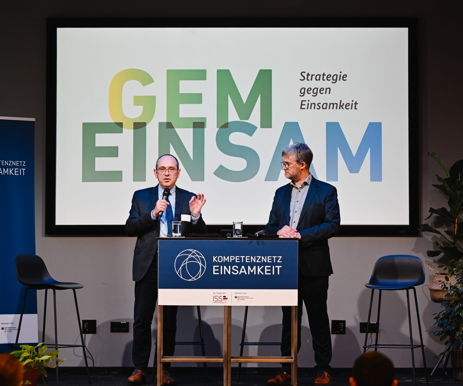 Zwei Personen stehen auf einer Bühne mit Mikrofonen und im Hintergrund sieht man den Schriftzug "GEMEINSAM - Strategie gegen Einsamkeit"