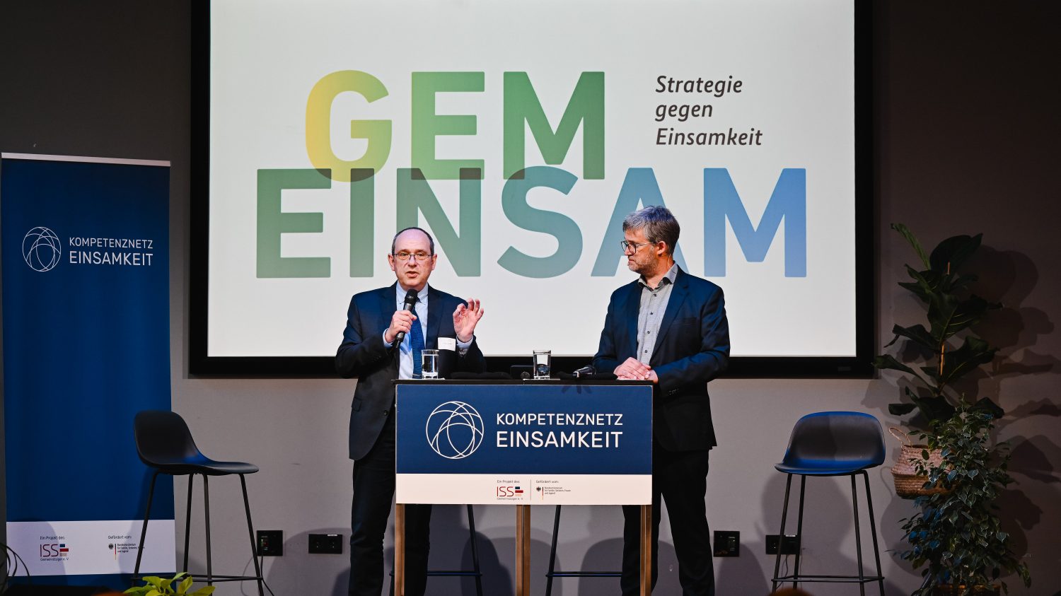 Zwei Personen stehen auf einer Bühne mit Mikrofonen und im Hintergrund sieht man den Schriftzug "GEMEINSAM - Strategie gegen Einsamkeit"