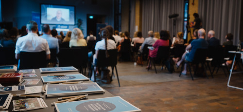 Das Publikum im Veranstaltungssaal ist von hinten fotografiert. Im Vordergrund ist ein Tisch mit Broschüren zu sehen.